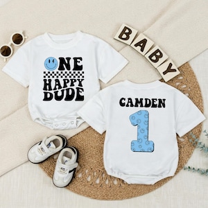 May include: Two white baby onesies with black text and graphics. One says "ONE HAPPY DUDE" with a smiley face and checkered pattern. The other says "CAMDEN 1" with a blue smiley face number one. White baby shoes and sunglasses are also in the image.