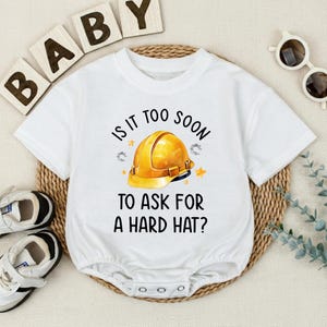 May include: White baby onesie with the text "IS IT TOO SOON TO ASK FOR A HARD HAT?" and a yellow hard hat graphic. The onesie has short sleeves and snaps at the bottom. The word "BABY" is spelled out in wooden blocks.