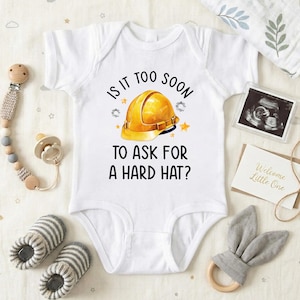 May include: White baby onesie with the text "IS IT TOO SOON TO ASK FOR A HARD HAT?" and a yellow construction helmet graphic. The onesie is surrounded by baby accessories, including a pacifier, booties, and a wooden teether.
