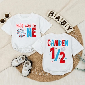 Könnte beinhalten: Zwei weiße kurzärmelige Babybodys mit roten und blauen Grafiken. Einer zeigt den Text Half way to ONE mit einem Feuerwerk-Symbol. Der andere zeigt den Namen CAMDEN mit der Zahl 1/2 und einem Feuerwerk-Symbol, dazu Babyschuhe.