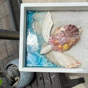 Könnte beinhalten: Ein gerahmtes Kunstwerk mit einer Meeresschildkröte aus Muscheln. Die Schildkröte besteht aus rosa, braunen und cremefarbenen Muscheln. Der Hintergrund zeigt einen sandigen Bereich und einen blauen Harz-Ozean, alles in einem weißen Rahmen.