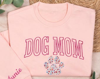 May include: Pink sweatshirt with the text "DOG MOM" in pink and white lettering. A floral paw print is below the text. The left sleeve has the name "Junie" embroidered in pink.
