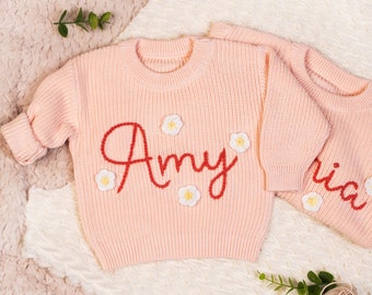 May include: Two pink knitted sweaters with embroidered white flowers and names. The sweater on the left says "Amy" and the sweater on the right says "Ria".