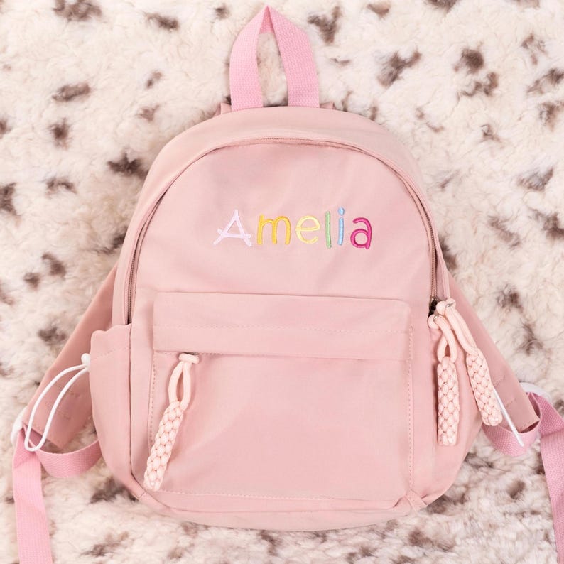 May include: A pink mini backpack with a rainbow embroidered name "Amelia" on the front. The backpack has two adjustable straps and a zippered pocket on the front.
