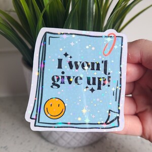 May include: A blue holographic sticker with a white border and a red paperclip. The sticker has a white background with a yellow smiley face and the text "I won't give up!" in black.