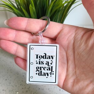 May include: A clear keychain with a white background and black text that reads "Today is a great day!" The text is surrounded by small black stars. The keychain has a silver ring.