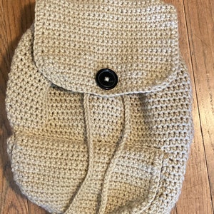 Puede incluir: Una mochila de crochet beige con cierre de solapa y un botón negro. La mochila tiene un cierre de cordón y un pequeño bolsillo en la parte delantera.