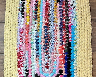 May include: An oval-shaped rag rug featuring a thick, solid pale yellow border surrounding a multi-colored center. The interior is composed of concentric loops of fabric strips in various patterns and colors, including pink, blue, red, black, and white, creating a textured, woven appearance.