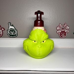Puede incluir: Un dispensador de jabón verde con temática de Grinch con una bomba marrón. El dispensador tiene una cara esculpida con ojos amarillos y una expresión gruñona. El dispensador está sobre una superficie blanca con decoraciones navideñas en el fondo.