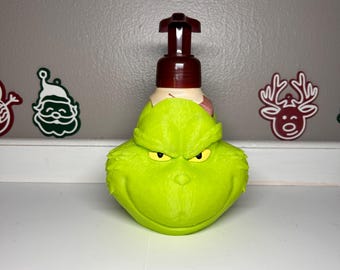 Peut inclure: Un distributeur de savon vert sur le thème du Grinch avec une pompe marron. Le distributeur a un visage sculpté avec des yeux jaunes et une expression grincheuse. Le distributeur est sur une surface blanche avec des décorations de Noël en arrière-plan.