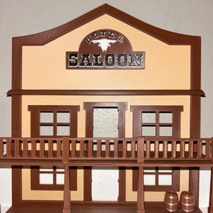 May include: A brown and tan miniature saloon with the word "SALOON" in silver lettering. The building has a balcony with a railing, windows, and two small barrels. The word "HOTEL" is above the word "SALOON".