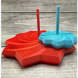 May include: Two 3D-printed geometric sculptures. One is a bright red starburst shape with a central vertical post. The other is a light blue starburst shape with a matching post. Both are on a light wood-grain surface.