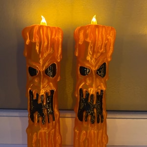 Könnte beinhalten: Zwei orangefarbene Halloween-Kerzen mit geschnitztem Gesichtsdesign. Die Kerzen haben schwarze Augen und Münder mit einem tropfenden Wachseffekt. Jede Kerze hat eine flackernde, warm getönte LED-Flamme.
