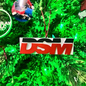 May include: Red and black Christmas ornament with the letters "DSM" in white. The ornament is hanging on a green Christmas tree.