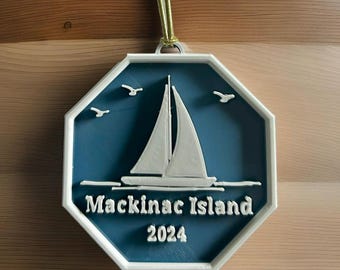 May include: White octagon-shaped ornament with a blue background. The ornament features a white sailboat with white birds flying above it. The text "Mackinac Island 2024" is printed in white on the ornament.