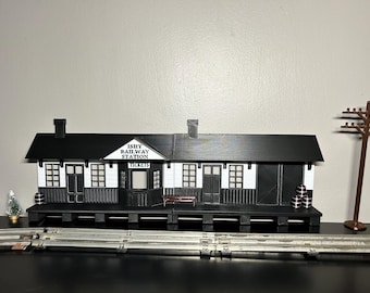 May include: A model railway station building in black and white with a sign reading ISHY RAILWAY STATION and TICKETS. The structure features a black roof, white walls, a ticket booth, a bench, and small barrels. A brown utility pole stands nearby.