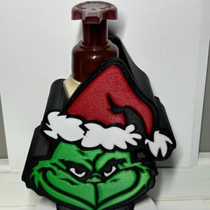 May include: A Christmas-themed soap dispenser featuring a green Grinch face with a red Santa hat and black accents. The dispenser has a brown pump and is designed to fit over a standard soap bottle.