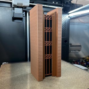 May include: A 3D printed architectural model of a tall building featuring two light brown textured side walls and a central dark brown section with a vertical grid pattern. The structure stands upright with a smooth finish and geometric details.