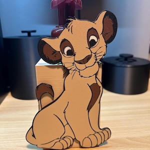 Pode incluir: Um recorte do personagem Simba de cor castanha clara de O Rei Leão, com detalhes castanhos nas orelhas, cauda e peito. O recorte está sobre uma superfície de madeira. A placa acima diz "SERVED HERE".