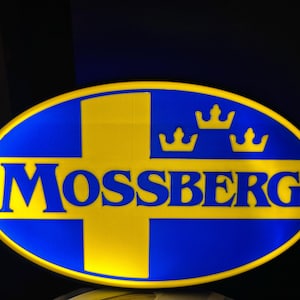May include: An oval sign with a blue background and a yellow cross and the word "MOSSBERG" in yellow. Three yellow crowns are above the word. The sign has a yellow border.