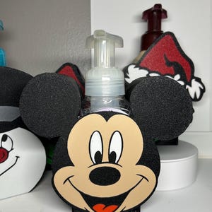 Puede incluir: Un dispensador de jabón de plástico transparente con un diseño de cara de Mickey Mouse. El dispensador tiene una base negra y orejas negras. La cara es de color canela con ojos negros, una nariz negra y una boca roja. Se ven otros dispensadores temáticos de personajes.