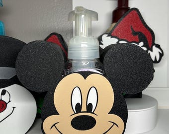 May include: A clear plastic soap dispenser with a Mickey Mouse face design. The dispenser has a black base and black ears. The face is tan with black eyes, a black nose, and a red mouth. Other character-themed dispensers are visible.