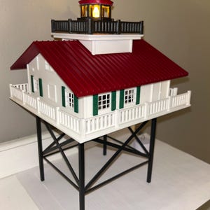 May include: A miniature lighthouse model with a red roof and a white body. The lighthouse has a black railing and a small, illuminated tower. The structure is supported by black metal legs and features green shutters.