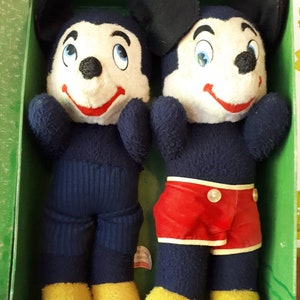 Puede incluir: Dos juguetes de peluche de Mickey Mouse en una caja verde. Un Mickey Mouse lleva pantalones azules y el otro lleva pantalones cortos rojos.