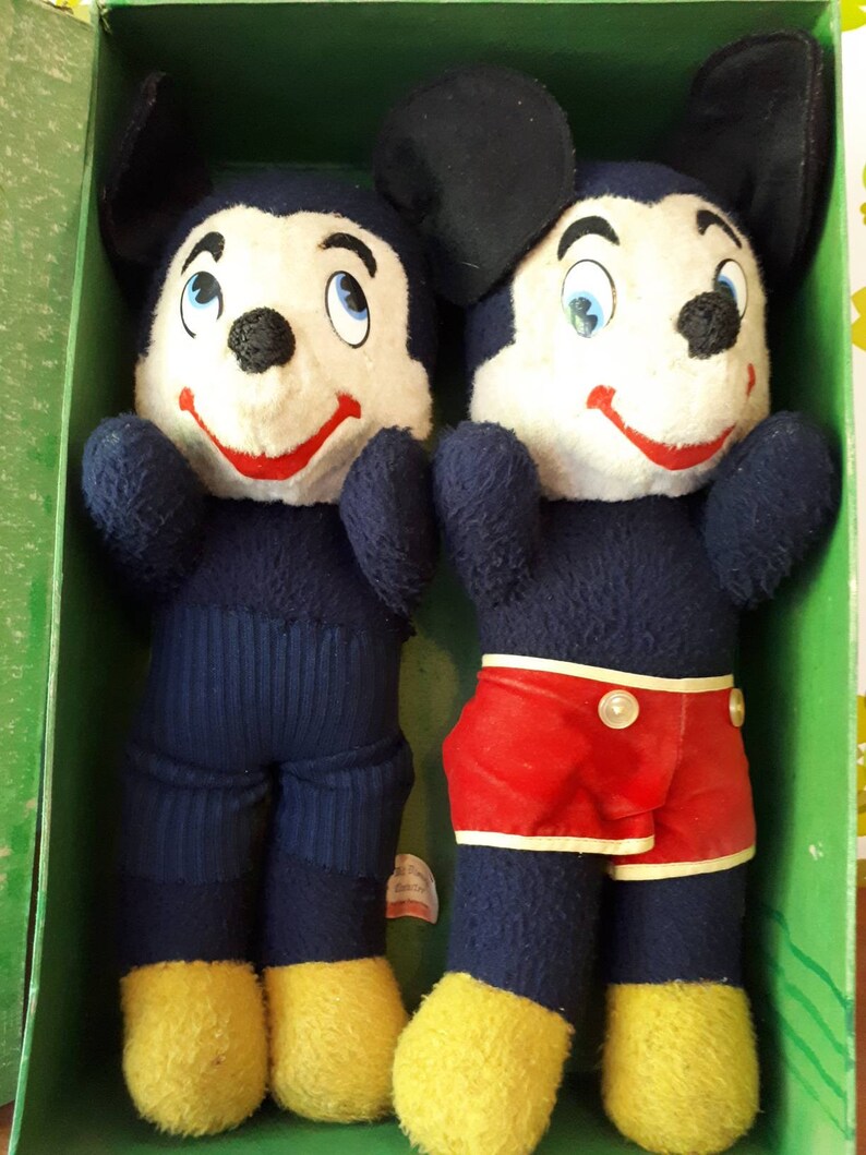 Puede incluir: Dos juguetes de peluche de Mickey Mouse en una caja verde. Un Mickey Mouse lleva pantalones azules y el otro lleva pantalones cortos rojos.