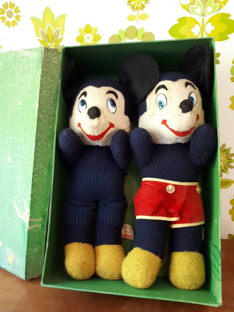 Puede incluir: Dos juguetes de peluche de Mickey Mouse en una caja verde. El Mickey Mouse de la izquierda lleva pantalones azules y zapatos amarillos. El Mickey Mouse de la derecha lleva pantalones cortos rojos y zapatos amarillos.