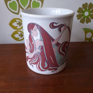 May include: White ceramic mug featuring an illustration of a red-haired mermaid. The mermaid has a green tail and is holding her long hair.