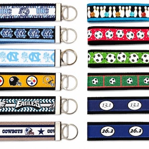 May include: A collection of twelve fabric keychains with metal hardware and split rings. The designs feature various sports themes including football team logos, soccer balls, bowling pins, and marathon distance markers labeled 13.1 and 26.2.