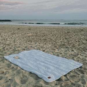 May include: A light blue quilted blanket with a rectangular shape, resting on a sandy beach. The blanket has a stitched, striped pattern and a small, rectangular tag in the corner. The ocean and sky are visible in the background.