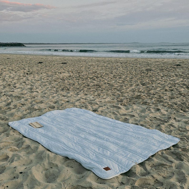 May include: A light blue quilted blanket with a rectangular shape, resting on a sandy beach. The blanket has a stitched, striped pattern and a small, rectangular tag in the corner. The ocean and sky are visible in the background.