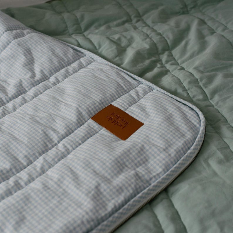 May include: Close-up of a quilted blanket with a light blue and white gingham pattern. The blanket has a brown leather tag with text and a zipper. The background is a solid sage green color. The blanket is likely for a bed or a baby.
