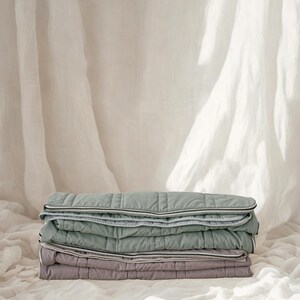 May include: A stack of three quilted blankets in shades of green and purple. Each blanket has a white and gray trim. The blankets are neatly folded and stacked on top of each other. The background is a soft, white fabric.