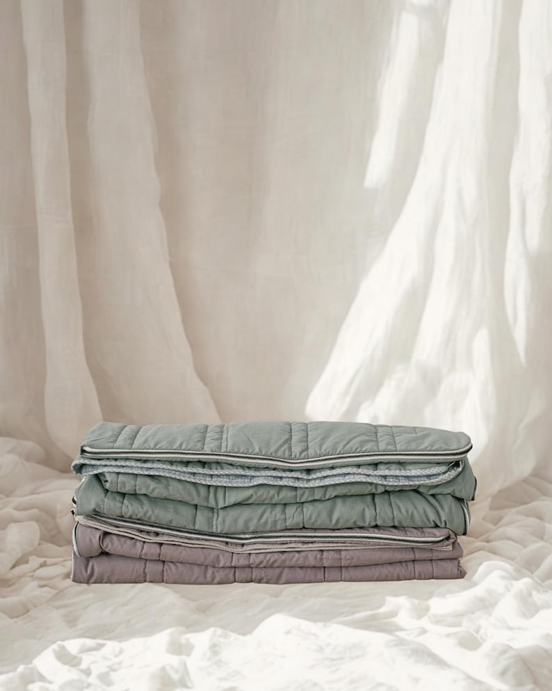 May include: A stack of three quilted blankets in shades of green and purple. Each blanket has a white and gray trim. The blankets are neatly folded and stacked on top of each other. The background is a soft, white fabric.