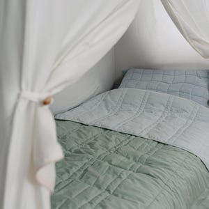 May include: A bed with a sage green quilted blanket and a light blue quilted pillow. A light blue blanket covers the top of the bed. White curtains are draped over the bed frame, tied with a wooden toggle.