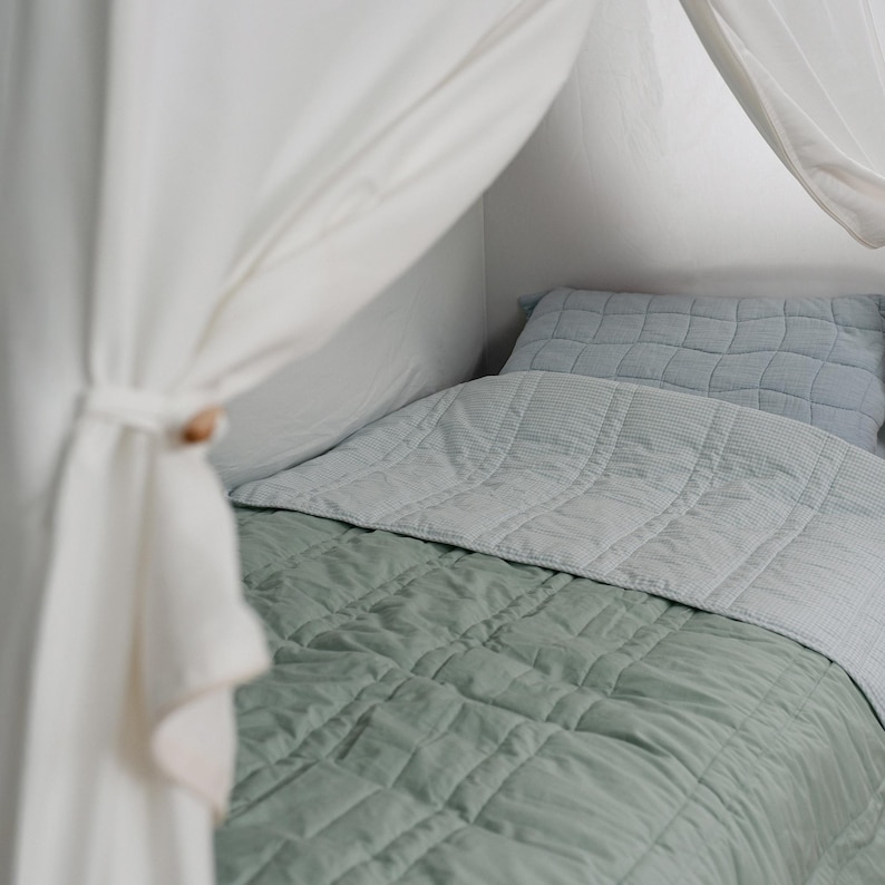 May include: A bed with a sage green quilted blanket and a light blue quilted pillow. A light blue blanket covers the top of the bed. White curtains are draped over the bed frame, tied with a wooden toggle.