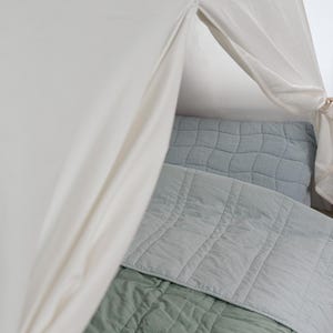 May include: A soft, off-white canopy frames a bed with a light blue quilted pillow and a matching blanket. A sage green blanket is visible at the bottom. The bedding has a textured, grid-like pattern.