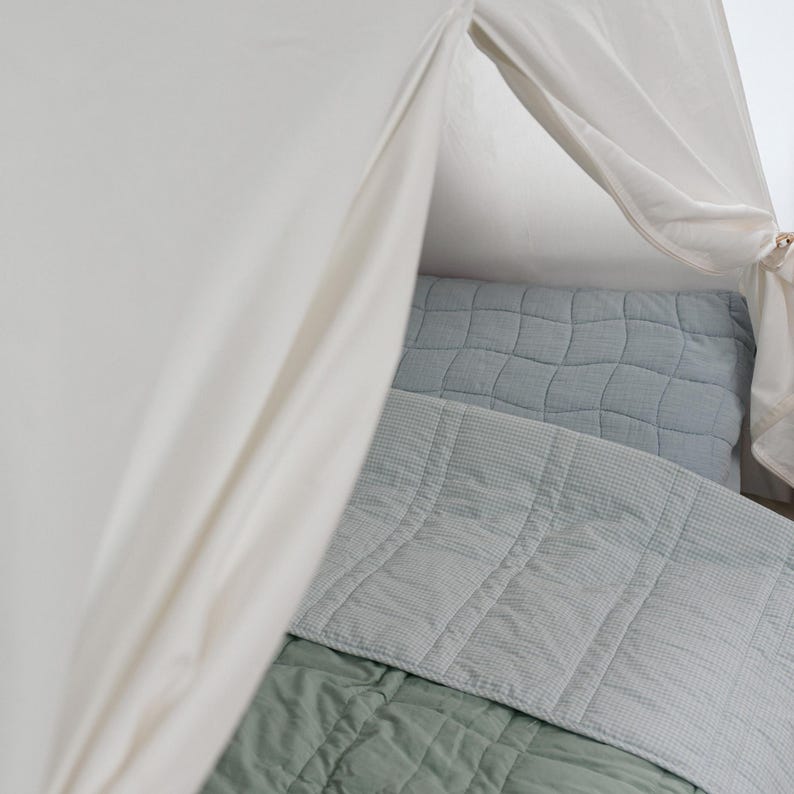 May include: A soft, off-white canopy frames a bed with a light blue quilted pillow and a matching blanket. A sage green blanket is visible at the bottom. The bedding has a textured, grid-like pattern.