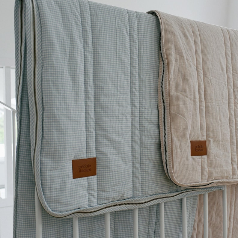 May include: Two baby quilts, one light blue with a gingham pattern and the other beige, are draped over a white crib. Both quilts have a quilted design and a brown leather tag that reads "pebble tacker".