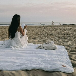 May include: A white and blue striped quilted picnic blanket is spread on a sandy beach. A light gray bag sits on the blanket. A person in a white dress sits on the blanket near the ocean. The scene is bathed in soft, natural light.