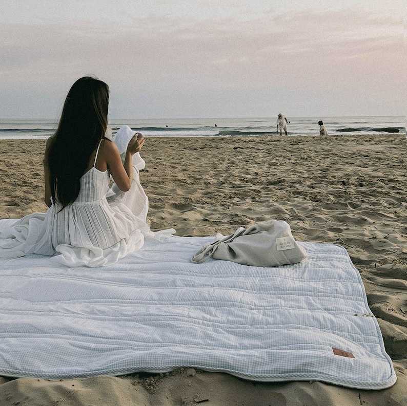May include: A white and blue striped quilted picnic blanket is spread on a sandy beach. A light gray bag sits on the blanket. A person in a white dress sits on the blanket near the ocean. The scene is bathed in soft, natural light.