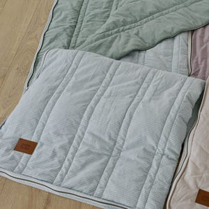 May include: A close-up of several quilted blankets in various pastel colors. The main blanket is light blue with a checkered pattern and a brown leather tag. Other blankets are sage green and pale pink. The blankets have a zipper closure.