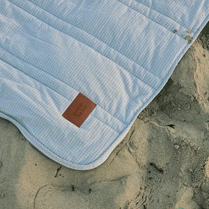 May include: A light blue and white gingham patterned blanket with a brown leather tag that reads "pebble sucker". The blanket is quilted with a zipper closure and is on a sandy surface.
