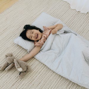 Peut inclure: Un enfant sourit, allongé sur un matelas de couchage à rayures bleu clair et blanc, avec un oreiller assorti. L'enfant tient un animal en peluche. Le matelas est posé sur un tapis beige.