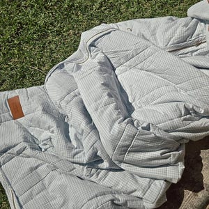 May include: A light blue and white gingham patterned quilted blanket or jacket. The item has a zipper and a brown leather tag. The item is laying on green grass.