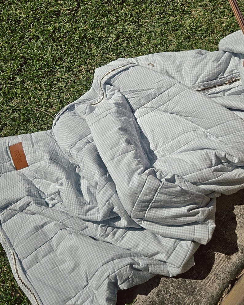 May include: A light blue and white gingham patterned quilted blanket or jacket. The item has a zipper and a brown leather tag. The item is laying on green grass.