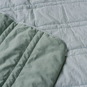 May include: A close-up of a quilted blanket with a light blue and white gingham pattern. The blanket has a solid sage green border with vertical stitching and a silver zipper.